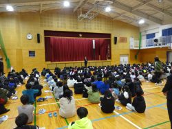 体育館できれいに整列して座っている子どもたちと、舞台下の中央に立っている先生を体育館の後方から写した写真