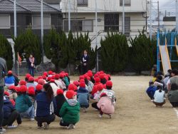 校庭で、整列してしゃがんでいる子どもたちと、子どもたちの前に立って話をしている様子の先生の写真