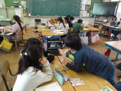 教室で、机を向かい合わせに付けて4名ずつほどのグループを作っており、タブレットを操作しながら話し合いを行っている子どもたちの写真