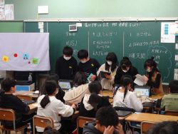 教室の黒板前に5名の生徒が立っており、手に持ったタブレットのようなものを操作している写真