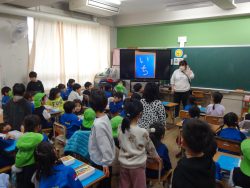 たくさんの子どもたちが集まっている教室内で、数名の子どもが緑色の帽子を被っており、正面に立つ先生の方を見ている写真