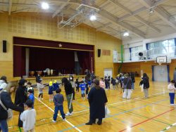 体育館内で、子どもたちが保護者と一緒に立ち、舞台の方を向いている写真