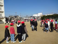 校庭に背の小さな子から大きな子まで一緒に並んでおり、一名の子が大縄を跳んでいる瞬間の写真