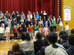 保護者の方々が見守る中、体育館に設置されたひな壇に並んで立っている子どもたちの写真