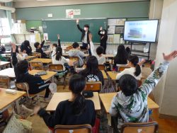 大きなモニターが用意された教室内で、黒板の前に立つ関係者や椅子に着席している子どもたちが手を挙げている写真
