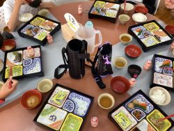 テーブル上にご飯や汁物、おかずの入った容器、お茶の入ったコップなどが並んでいる写真