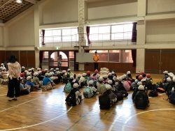 体育館に、帽子姿でリュックを背負った子どもたちが、並んで座っている写真