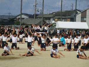 運動場の中心で円陣を組んで肩車をし、上に乗っている児童が手をつないで背中をそらし、その周りを膝をつき上半身を立たせ手を挙げた児童や、足を延ばして座り、手を左に伸ばしてる児童の演技をしている写真