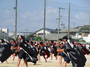運動場で裸足になり、黒の法被を着て、赤の鉢巻と白の鉢巻をした児童が踊っている写真