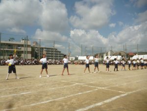 運動場に横一列に並んだ児童が足を肩幅ほどに開き、右手は腰、左手はまっすぐ伸ばしている様子の写真