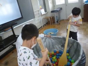 大きな鍋の模型にカラフルなボールが入れられ、鍋の回りで大きなしゃもじを持って児童2人が混ぜている写真