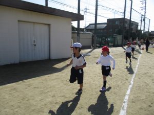 晴天の下、体操服姿で赤や白色の帽子を被った子供たちが運動場の周りを一生懸命走っている様子を写した写真