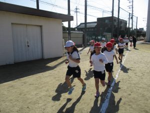晴天の下、体操服姿で赤や白色の帽子を被った子供たちが運動場の周りを走っている様子の写真