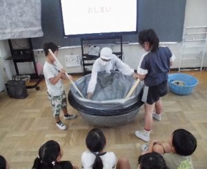 白いフードと白衣を着た1名が大きな鍋の模型を押さえており、両脇に立っている2名の児童が、しゃもじで鍋を混ぜている写真