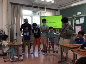 映像の写し出された大型モニターの前に4名の児童と2名の先生、机の自席に座っている子ども達が写っている和やかな授業風景の写真