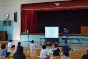 体育館の舞台下に大きなモニターが用意され、映し出された資料を見ながら話している様子の関係者と、床に座ってモニターの方を向いている生徒たちの写真