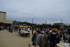 エプロンや三角巾を身に着けた人たちが校庭に集まっており、数か所に設置されたテーブルの周りに立って作業をしている写真