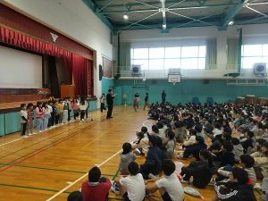 体育館にたくさんの生徒たちが集まって座っており、舞台の前に並んで立っている生徒や先生の方を向いている写真