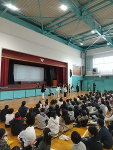 たくさんの生徒たちが座っている体育館で、舞台の前に立っている生徒が向かい合っている写真