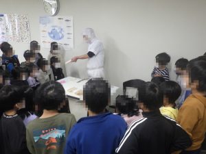 白衣を着た関係者がかまぼこを置いた台の後ろに立っており、周りに集まっている生徒たちへ説明をしている様子の写真