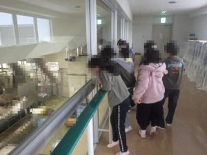 工場の内部が見える窓の前に立って覗き込むように見学をしている生徒たちの写真