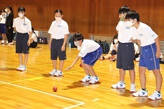 体育館で生徒5名が横一列に並んでおり、真ん中にいる生徒が赤いボールを転がし、周りの生徒がボールの動きを見守っているボッチャ体験の様子の写真