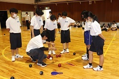 赤色や紺色のボールがいくつも転がっている場所に先生がかがんでおり、その周りに生徒6名が集まって先生の様子を見ているボッチャ体験の様子の写真
