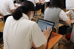 席に座っている女子生徒がタブレット端末を指で操作し、学習している様子を後方から写した写真
