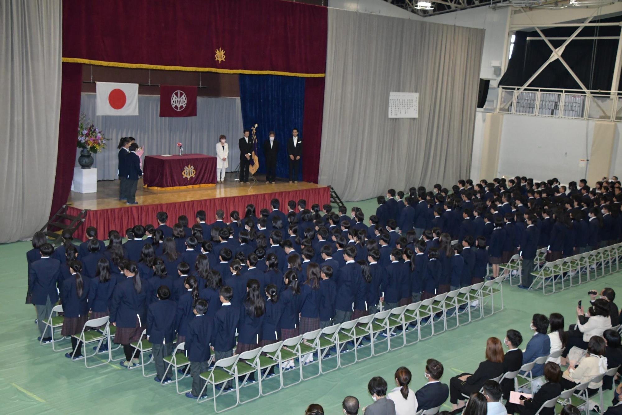 国旗と学校旗が飾られた体育館のステージ右側に4名の教師、左側に4名の生徒が並んで立ち、新入生たちが椅子から立ち上がっている入学式の様子の写真