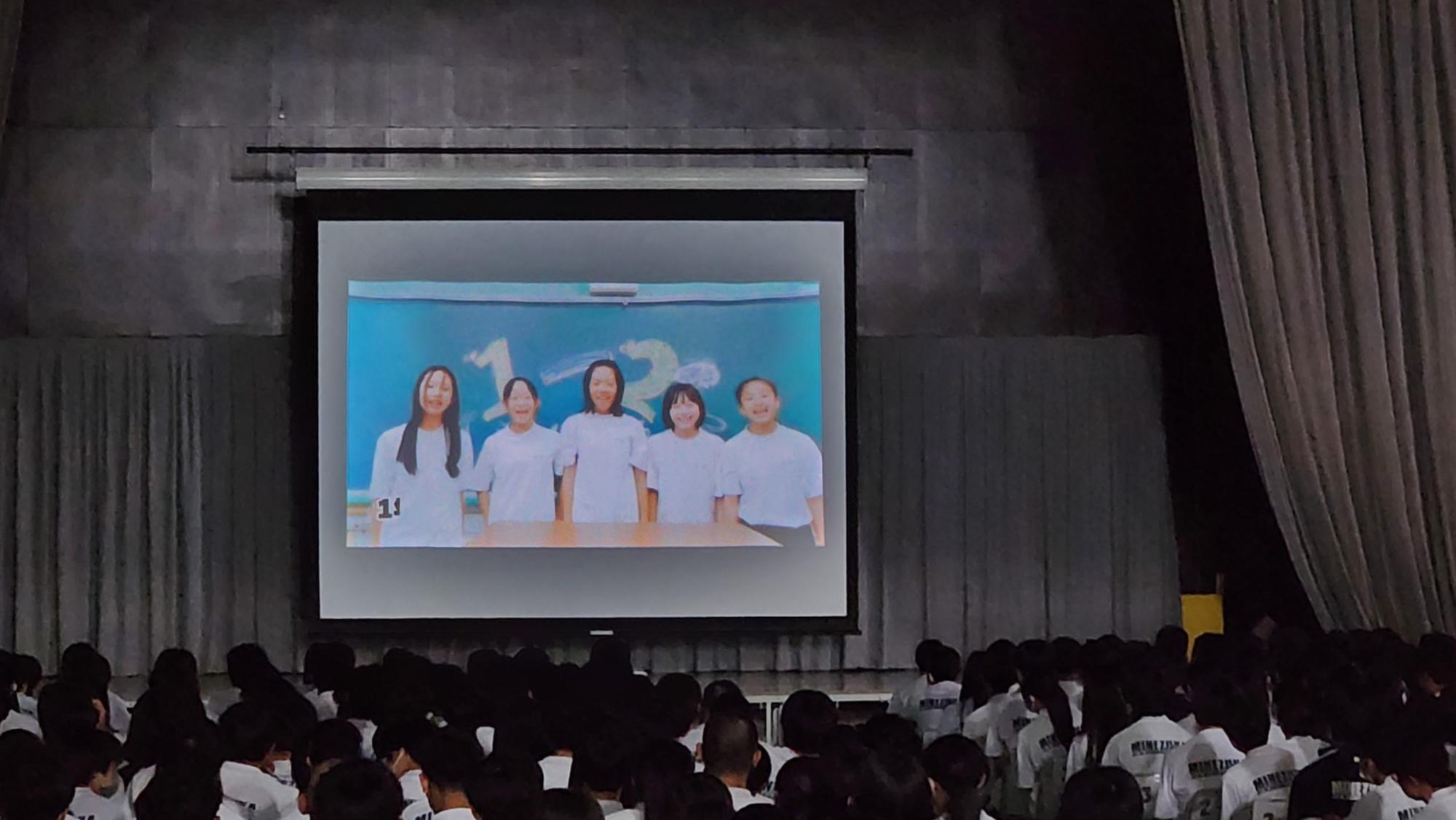 照明をおとし薄暗くなった体育館で、ステージに設置されたスクリーンに5名の女子生徒が並んでいる映像を体操着姿の生徒たちが見ている後ろ姿の写真