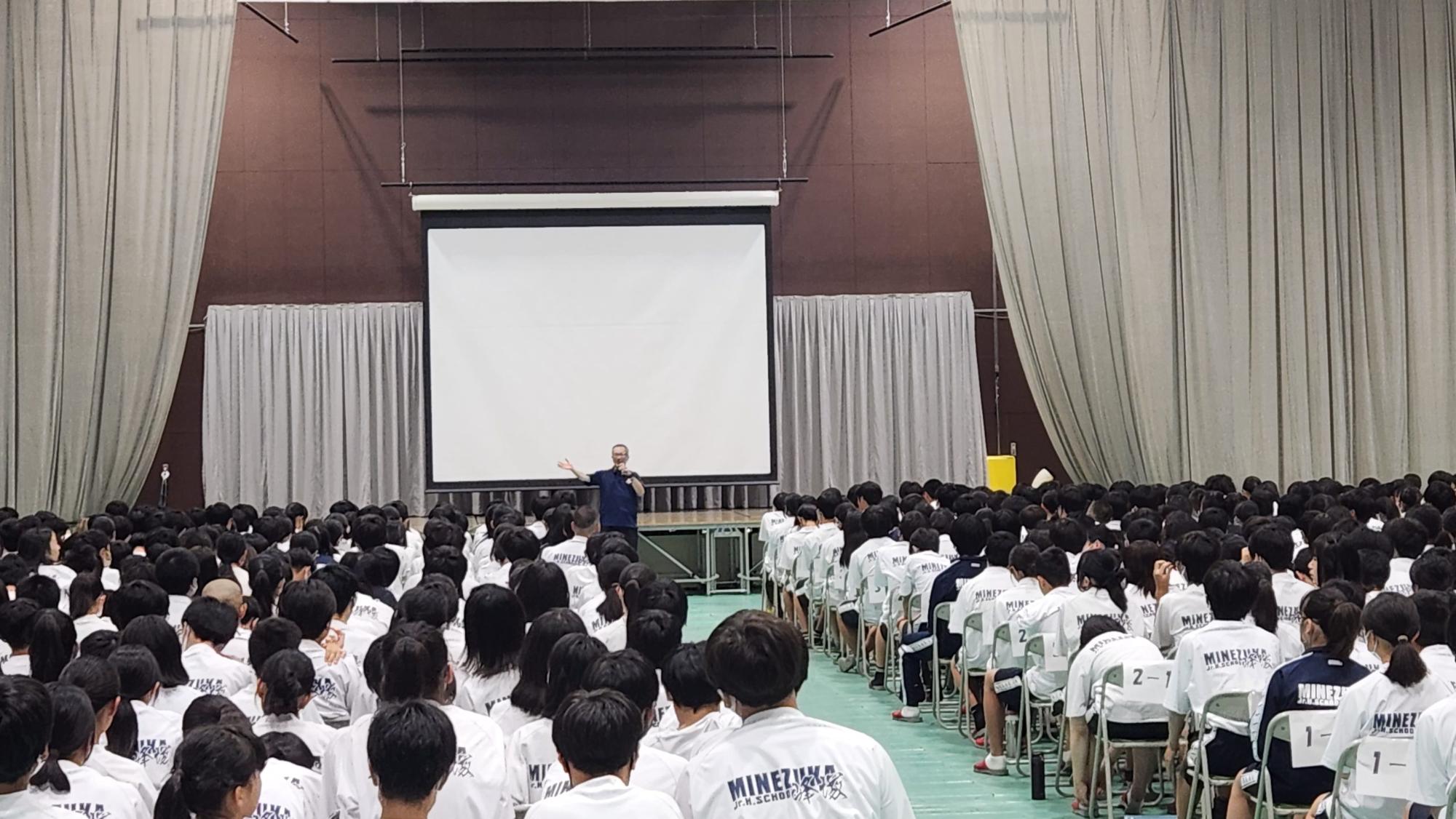 天井から吊るされた大きなスクリーンが設置されたステージの前に立ちマイクを持っている男性の話を、クラス別に座った体操着姿の生徒たちが聞いている後ろ姿の写真