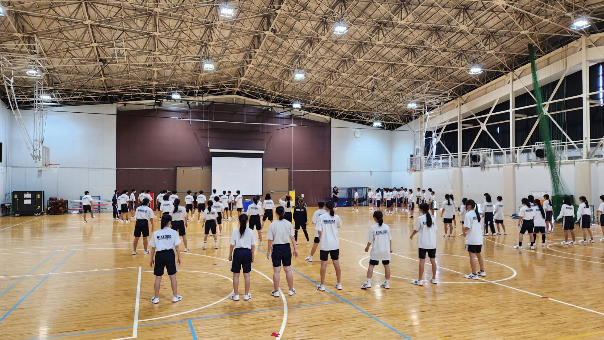 体育館で3年生の生徒たちが間隔をあけて左右に列をなして並びダンスの練習を行っている様子を後方から撮影した写真