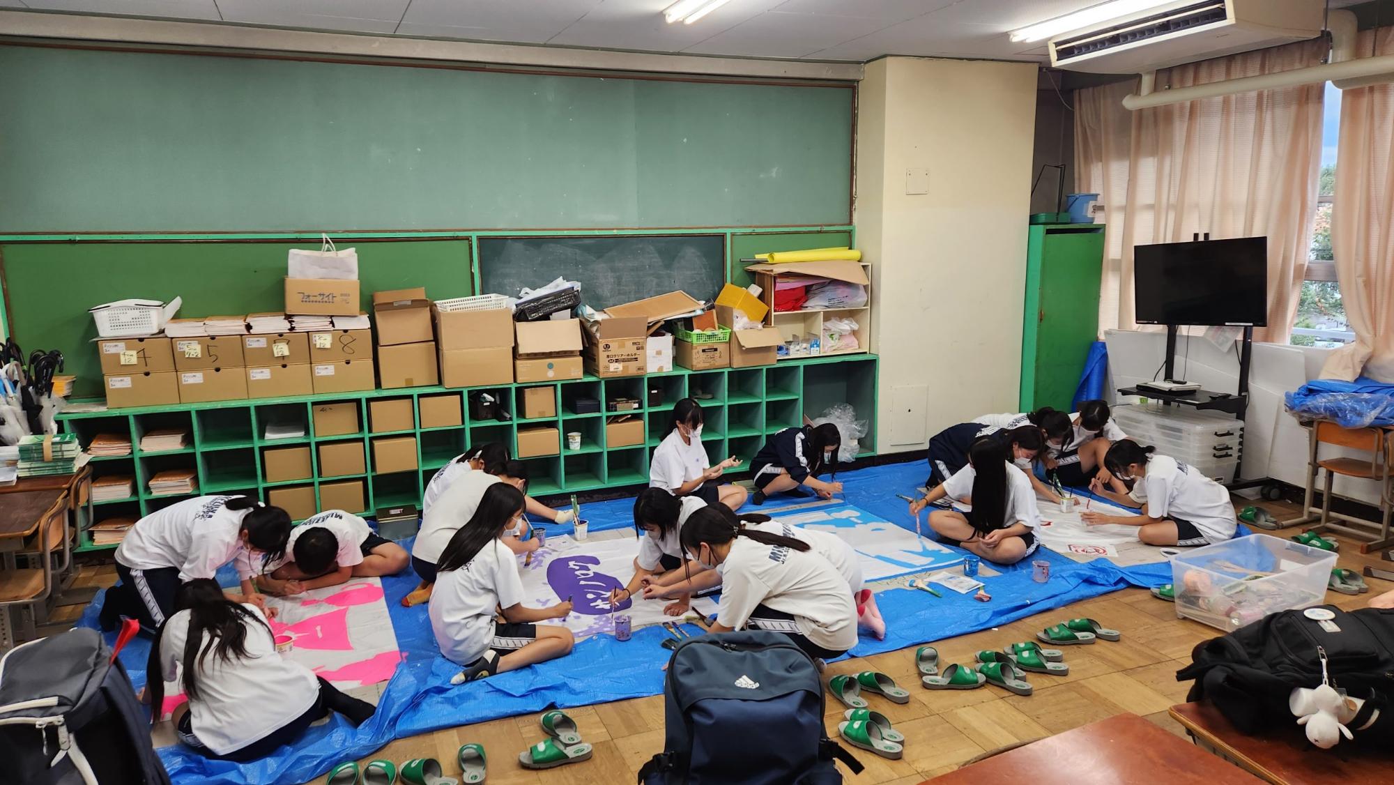 教室の後方の床にビニールシートを敷き、体操着姿の女子生徒が4枚の白旗にピンク色や水色などで色を塗っている学級旗づくりが行われている様子の写真