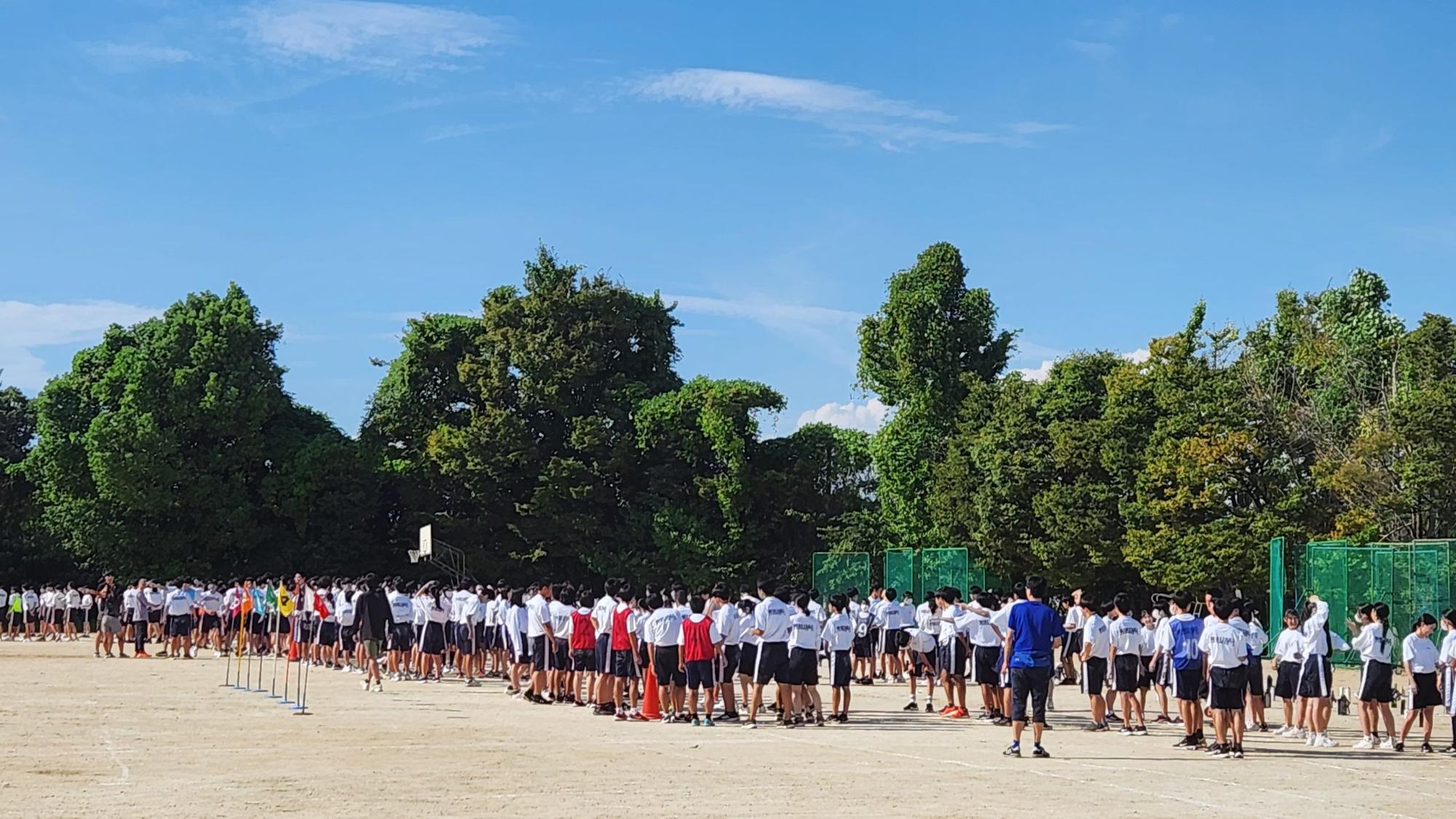学校の敷地内を囲むように木々が鬱蒼と茂った運動場の片隅に、体操着姿の3年生の生徒たちが集まっているリハーサルの様子の写真