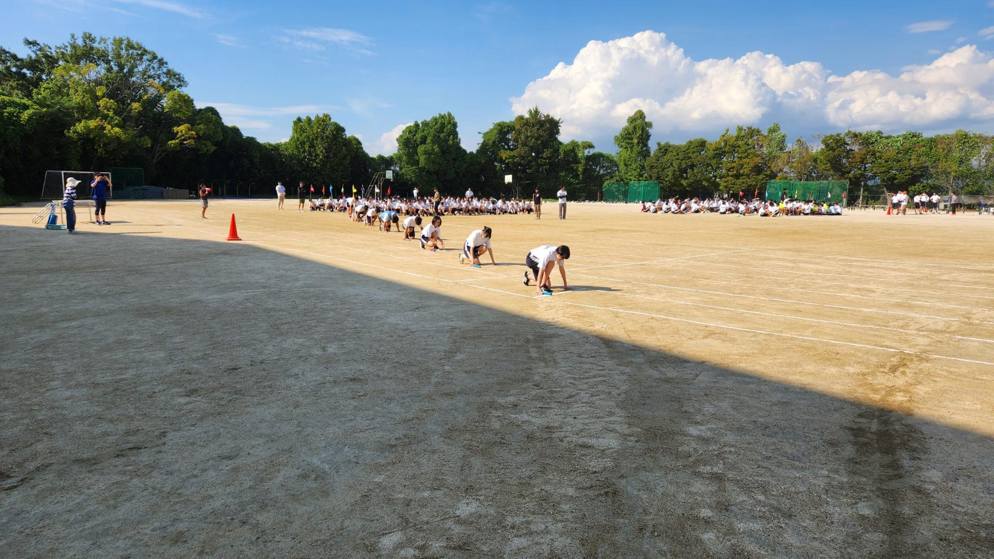 運動場に3年生が2か所に固まって集まり、8名の女子生徒がクラウチングスタートのポーズをとっている全員リレーの練習の様子の写真
