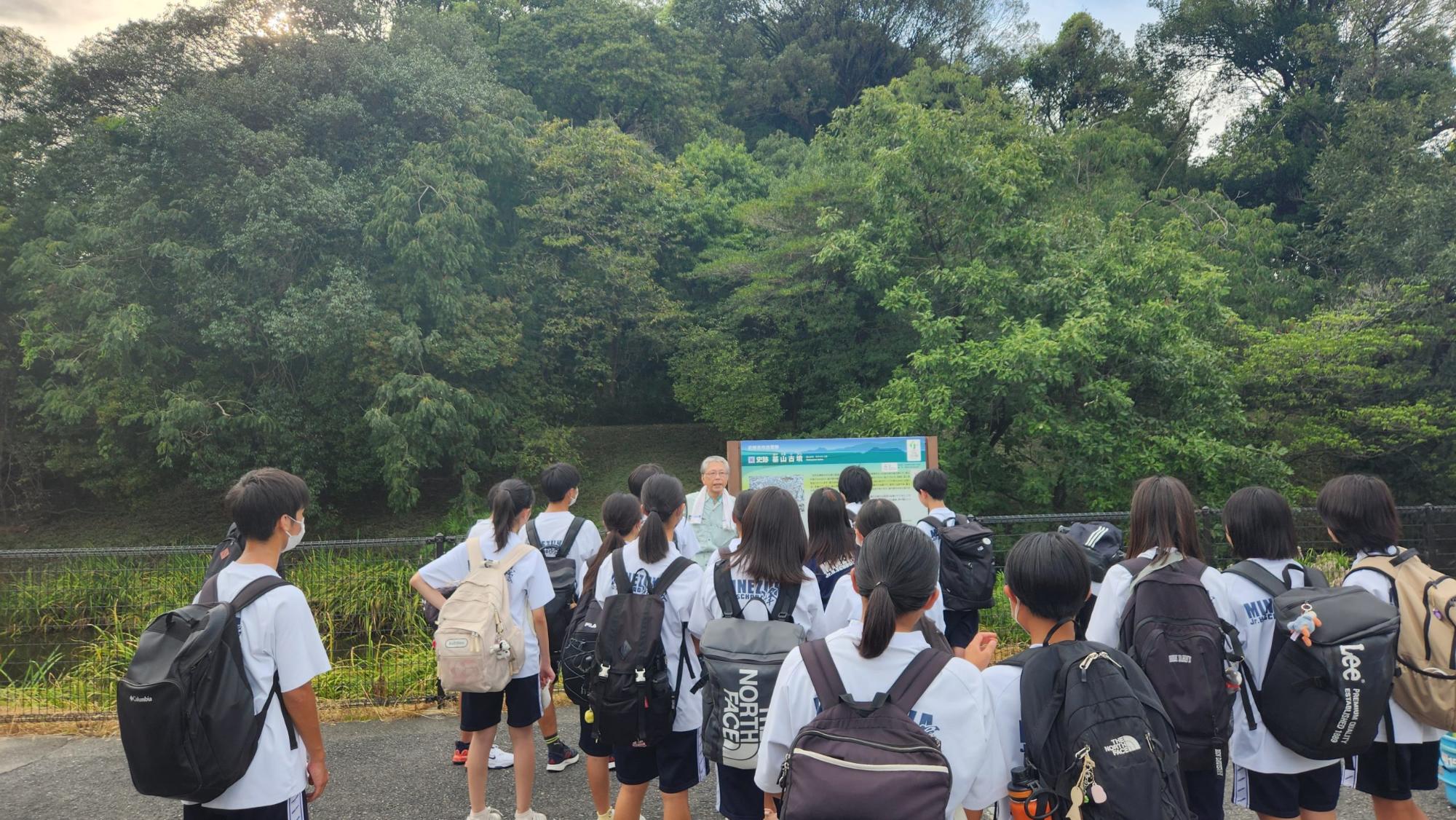 鬱蒼と茂った木々に囲まれた古墳の説明版の前で、作業着姿の男性の話をリュックを背負った生徒たちが聞いている様子の写真