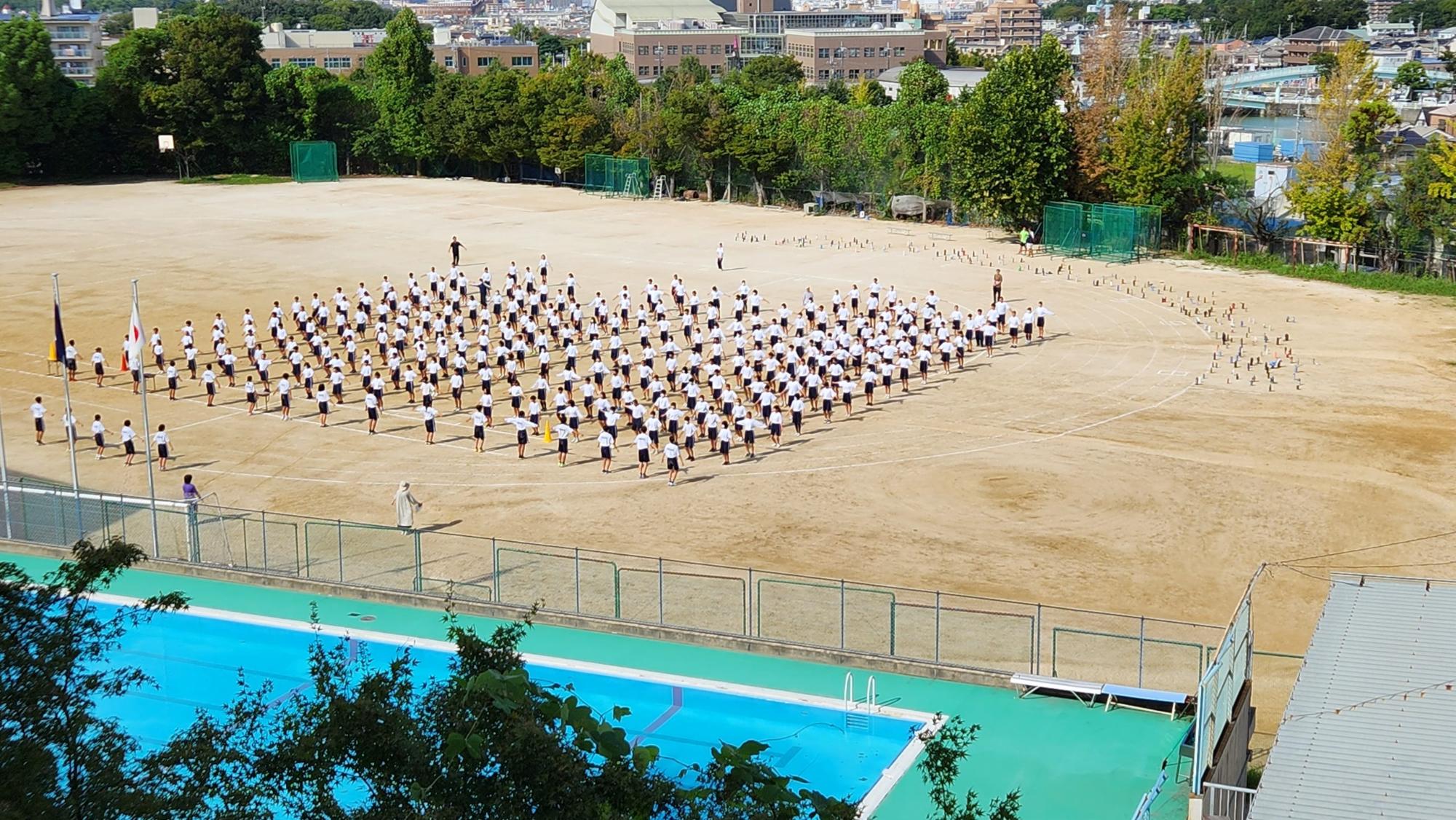 運動場の右半分に2年生が間隔をあけて立ち、ラジオ体操の練習が行われている様子の写真