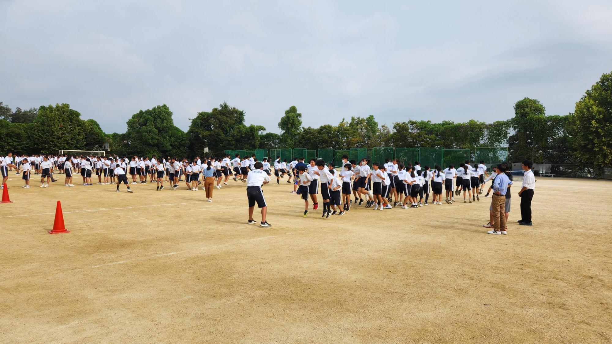 木々に囲まれた学校の運動場で教師が見守るなか、体操着姿の生徒たちが各クラスに分かれて大縄跳びの練習を行っている様子の写真