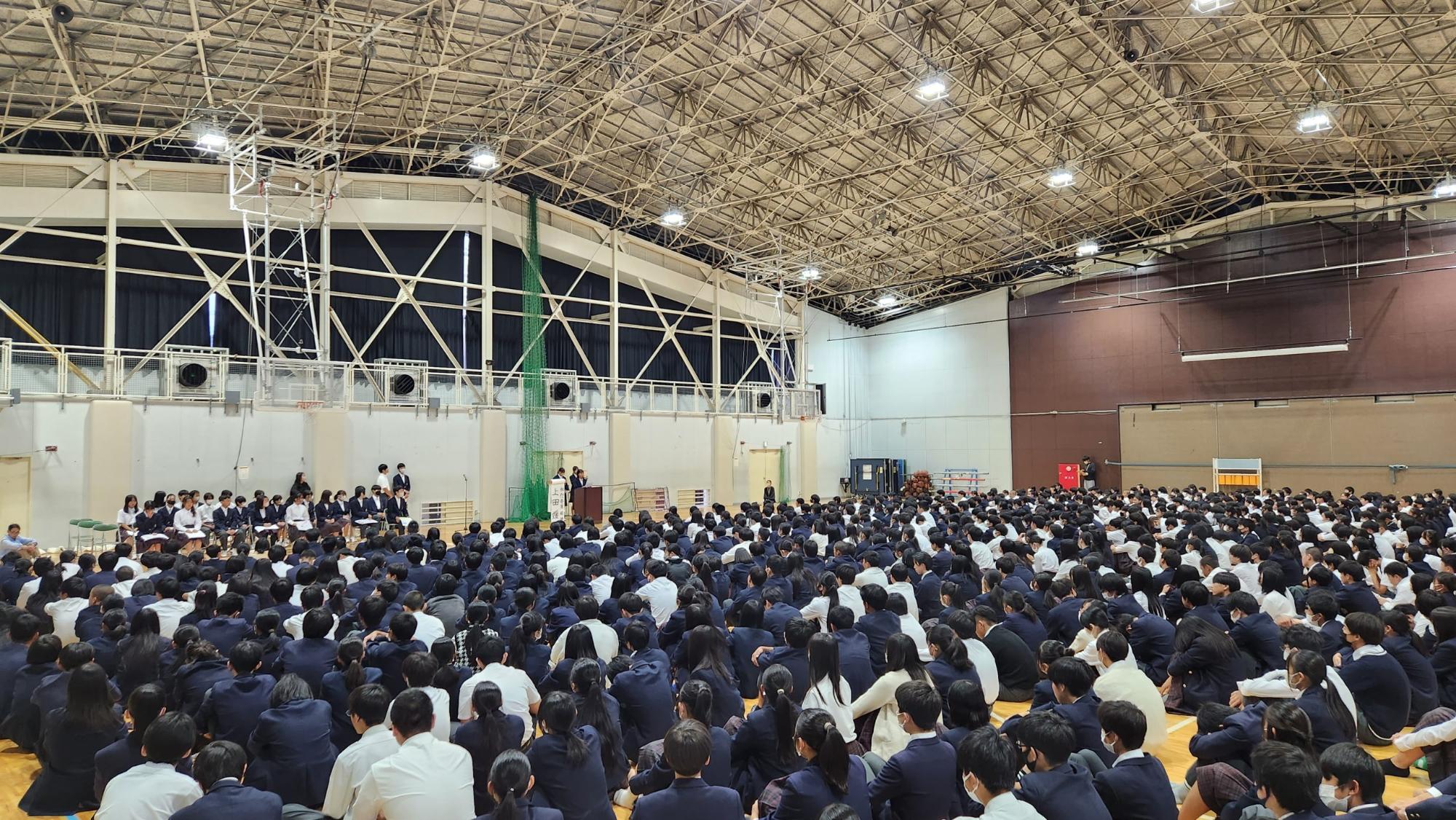 体育館の前方に立候補者名が書かれためくり台と演壇で立候補者が演説を行い、左側に設置されたパイプ椅子に立候補者が座り、体育館の床に体育座りをした全校生徒が座って聞いている立会演説会の様子の写真