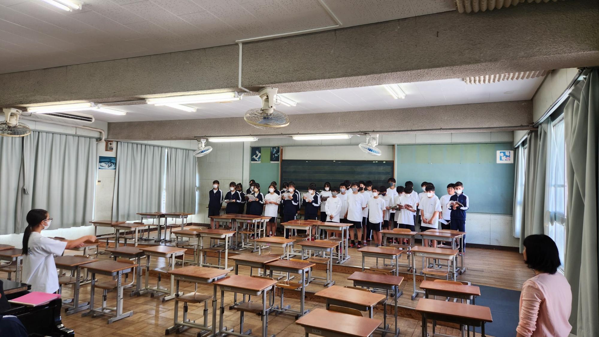 音楽室の後方に体操服やジャージ姿の生徒たちが立ち、女子生徒の式とピアノの伴奏に合わせて合唱の練習を行っている様子を、右側で女性教師が見ている様子の写真