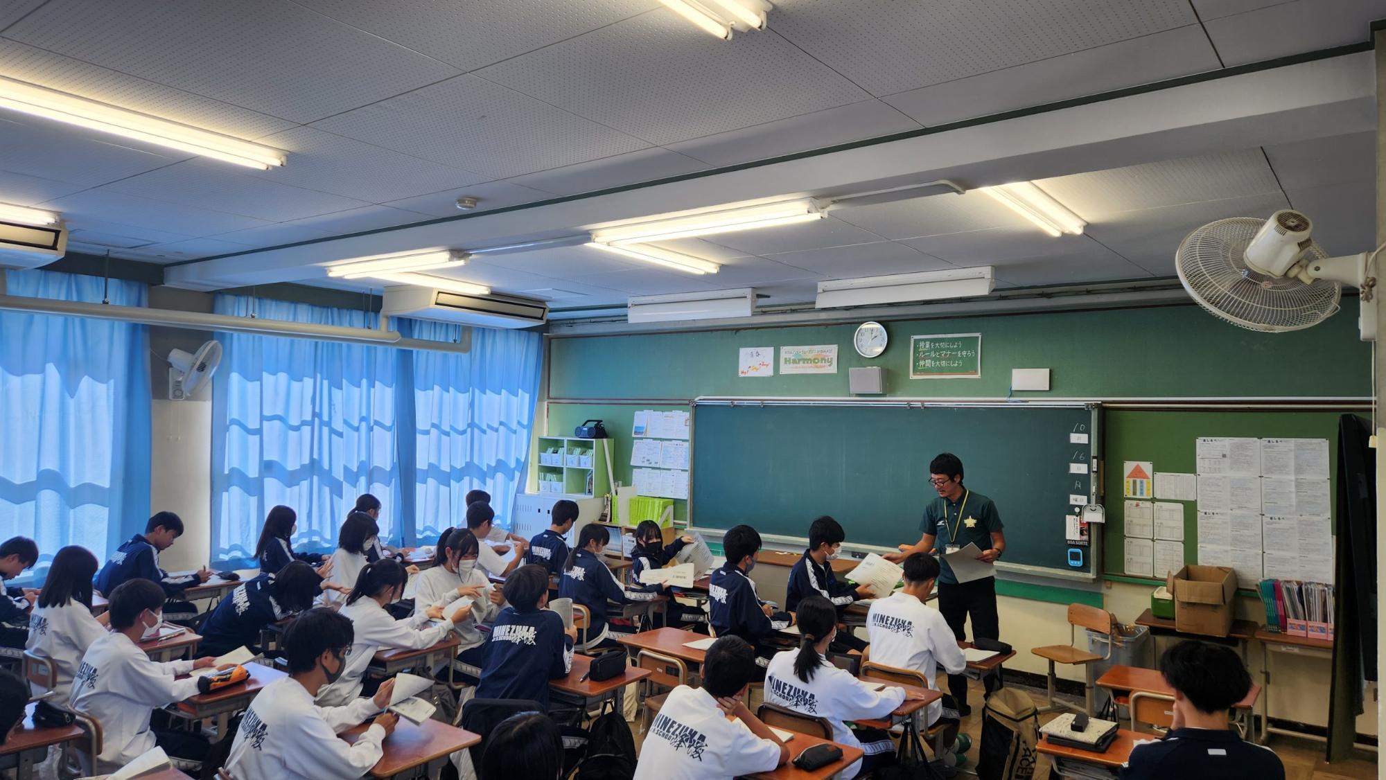 教室内で自席に座った生徒たちが、前の席から配られた資料を見たり冊子を綴じている学活の様子の写真