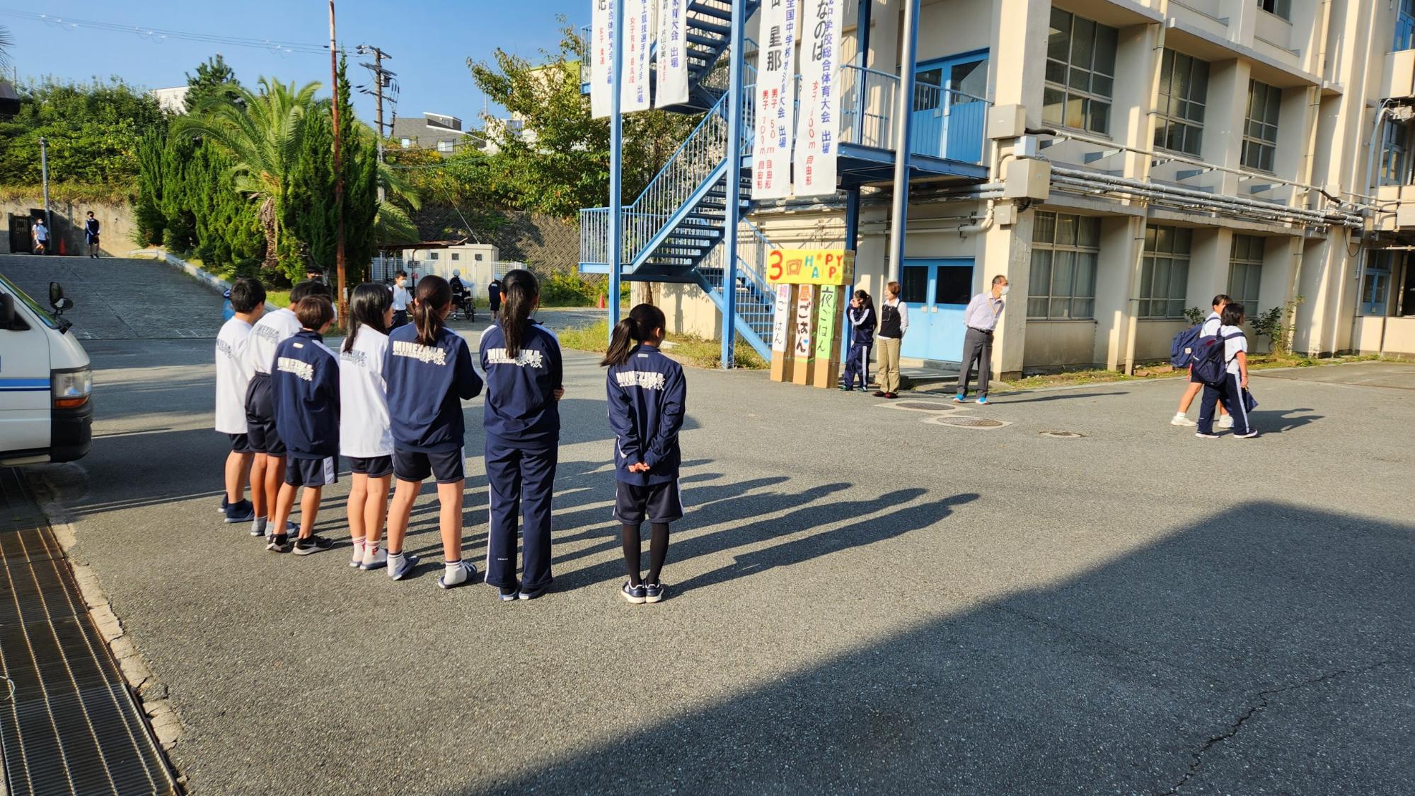 体操服やジャージ姿の7名の生徒たちと2名の教師と1名の女子生徒が校門の前で向かい合って立ち、あいさつ運動を行っている様子の写真