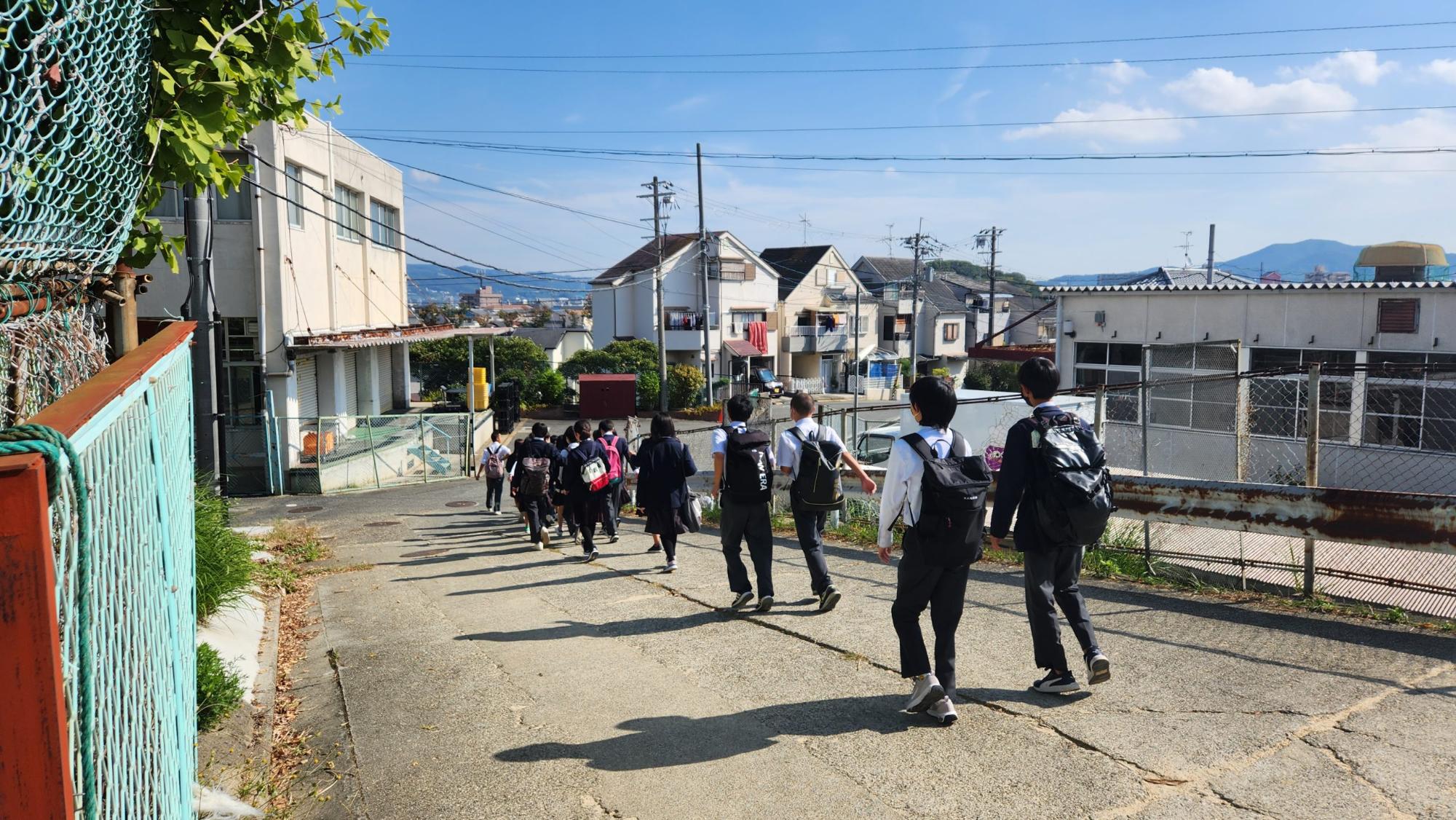 リュックを背負った2名ずつ並んだ生徒たちが学校を出発して行く様子の写真