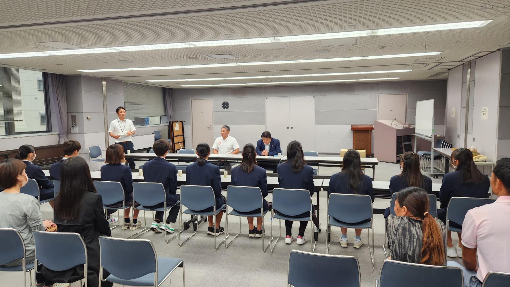 中央に並んで座っている市長・教育長、11名の生徒と関係者が向かい合って座り、左側に立っている男性が話をしている表敬訪問の様子の写真
