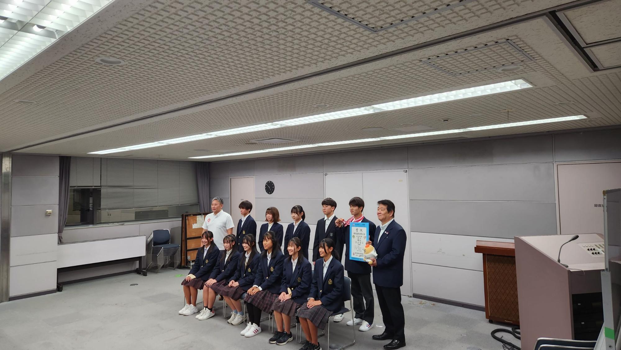 前列に6名の生徒、後列に5名の生徒と市長と教育長が立ち、左側に立つ男子生徒がメダルを首にかけ、右側の男性が賞状とぬいぐるみを持っている記念写真