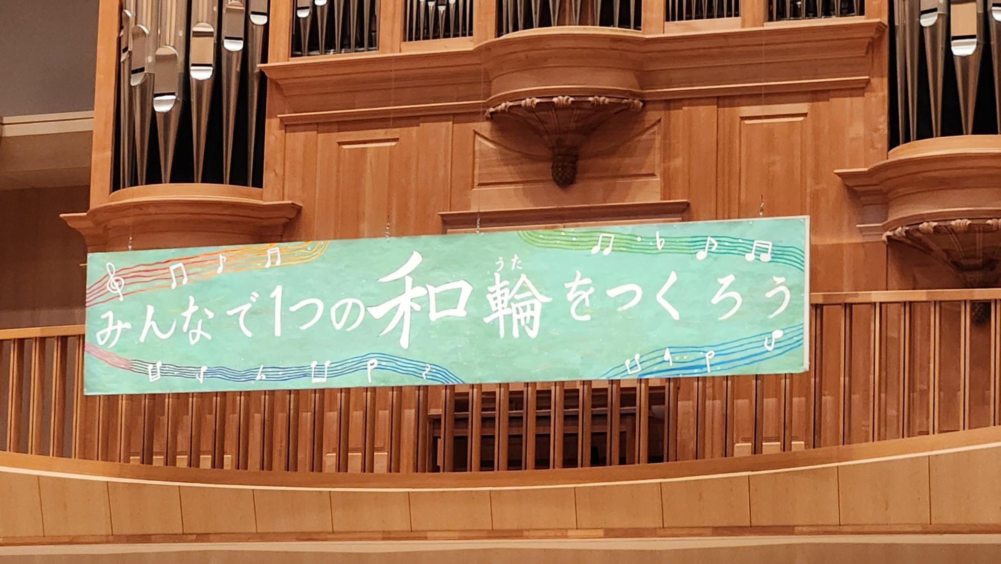 音符のイラストと「みんなで1つの和輪（うた）をつくろう」の文字が書かれた、ステージ上に設置された舞台看板の写真
