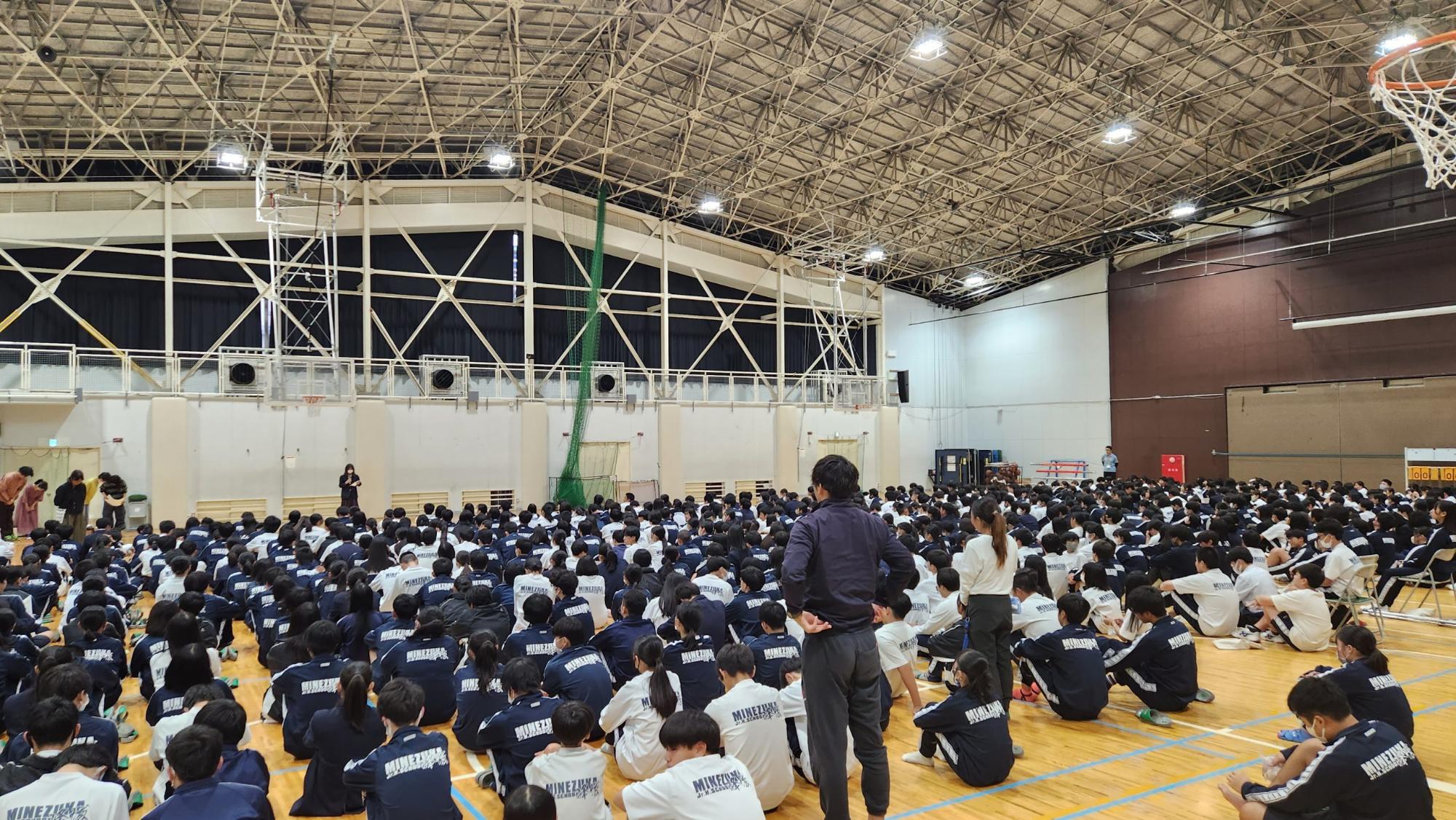 体育館の床に体育座りをしたジャージ姿の生徒たちが並んで座り、奥の壁側の中央に女子生徒が立ち、左前に4名の女性が生徒に向かってお辞儀をしている生徒総会の様子の写真