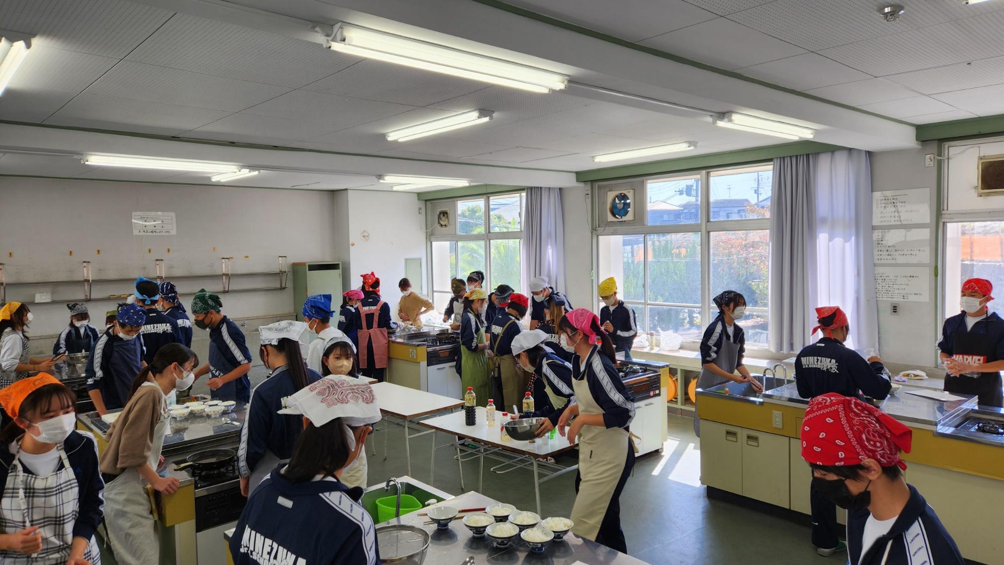 調理実習室で頭に三角巾をつけエプロンを着用した生徒たちがグループごとに分かれて調理を行っている様子の写真