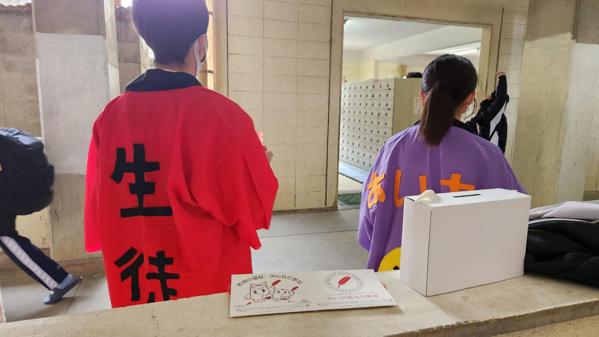 背中に黒文字で生徒会と書かれた赤色の法被を着た男子生徒と、背中にオレンジ色の文字であいさつと書かれた紫色の法被を着た女子生徒が靴箱の方向を向いて並んで立っている後ろ姿の写真