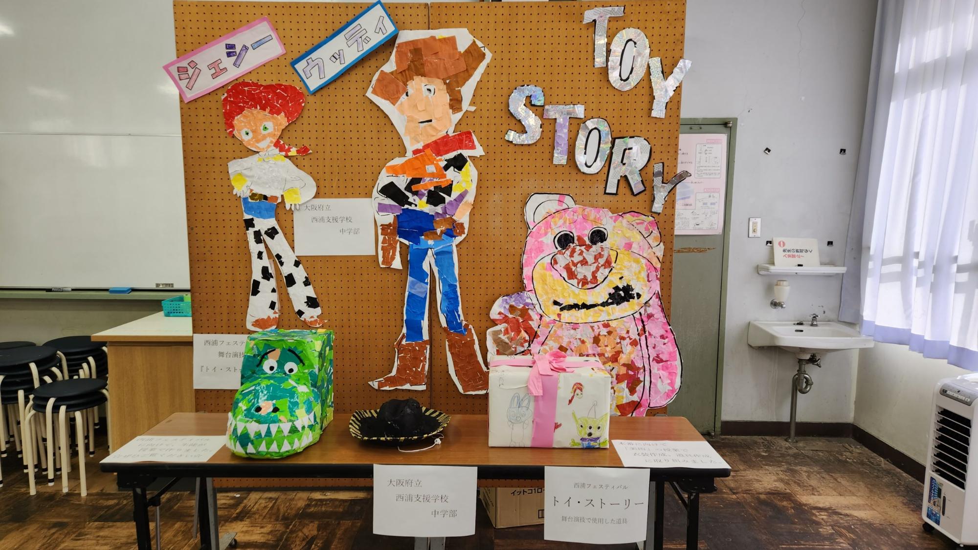 木製の掲示ボードにトイストーリーの主要キャラクターがちぎり絵で展示され、手前の長机にピンクのリボンとトイストーリーのイラストが描かれた包装紙でくるまれたで包まれたプレゼントの箱や黒色のテンガロンハットなどが並んでいる西浦支援学校の生徒が作成した作品の写真